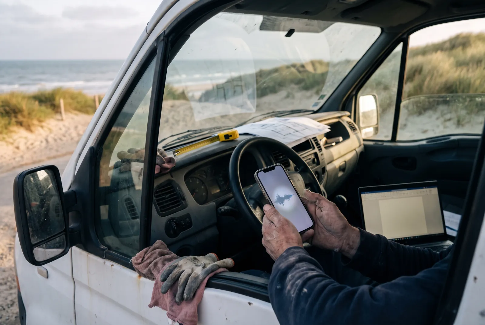 Les mains d'un artisan tiennent un téléphone en enregistrement vocal dans sa camionnette garée près des dunes